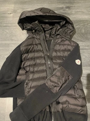 Svart dunväst med huva från Moncler - Snygg svart dunväst från Moncler med stickade ärmar och huva. Västen har dragkedja framtill, två sidofickor och ribbade muddar. Klassisk Moncler-logga på ärmen och quiltad design på fram- och baksidan. Perfekt för lager-på-lager och streetstyle. (Har fler bilder skriv vid intresse 