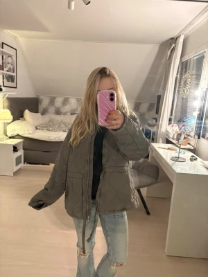 Olivgrön quiltad jacka med dragsko - Säljer en olivgrön quiltad jacka  i storlek S med dragkedja och dragsko i midjan från H&M, men liknar den populära jackan från Zara. Jackan har två stora fickor framtill och ett mönster av vågiga sömmar. Perfekt för lager på lager och en avslappnad stil. Prislappen är dessutom kvar.