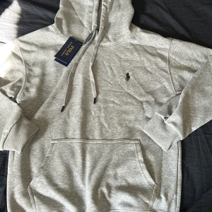 Grå Polo Ralph Lauren hoodie  - Säljer en stilren grå hoodie från Polo Ralph Lauren med klassisk broderad logga på bröstet. Tröjan har huva med dragsko och en stor magficka. Perfekt för en avslappnad och sportig look. Storlek S men passar bättre XS.