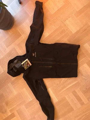 Svart Arc'teryx Gore-Tex regnjacka - Säljer en svart regnjacka från Arc'teryx med GORE-TEX-material. Jackan har huva, hel dragkedja framtill och två bröstfickor med dragkedja. Perfekt för blöta dagar och outdoor-äventyr. Snygg och funktionell design med diskret logga på bröstet. Grym vattentäthet 