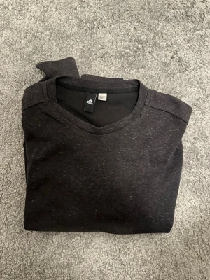 Mörkgrå hoodie från Adidas - Mörkgrå hoodie från Adidas med klassisk känguruficka framtill och långa ärmar. Tröjan har en enkel design utan tryck och är tillverkad i mjukt material som känns skönt mot huden. Perfekt för en avslappnad och sportig stil.