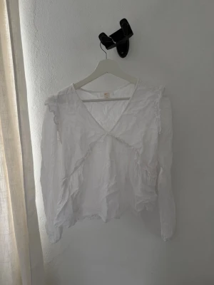 Vit blus med volang från Gina Tricot - Söt vit blus från Gina Tricot med v-ringning och volangdetaljer längs framsidan. Blusen har långa ärmar och är tillverkad i ett lätt, tunt tyg som ger en luftig känsla. Perfekt att styla med jeans eller kjol för en trendig look.