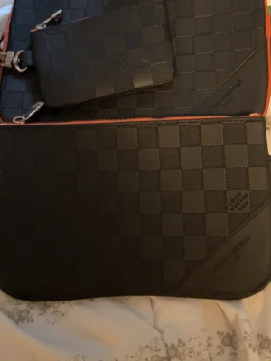  svart rutmönster axelväska - Axelväska i svart med klassiskt rutmönster och orange detaljer. Väskan har en matchande mindre väska/ficka med dragkedja och axelrem. Tillverkad i tåligt läder med präglad logotyp på framsidan.