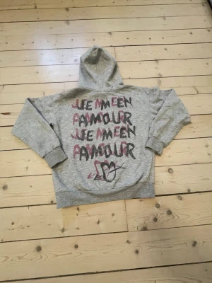 Grå hoodie med tryck 'Je m'en amour' - Grå hoodie med huva och ribbade muddar. På bröstet står det 'Je m'en amour' i röd och svart text, och på ryggen finns samma text i större format flera gånger samt ett hjärta. Mjuk bomullskänsla och avslappnad passform.
