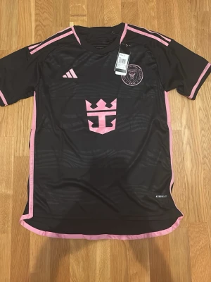 Svart Inter Miami matchtröja Adidas - Helt ny oanvänd Inter Miami tröja med rosa detaljer och klubbmärke på bröstet. Tröjan har korta ärmar, rund hals och rosa ränder på axlarna. Tillverkad i lätt och ventilerande material.