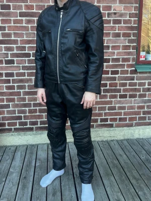 Svart skinnjacka med dragkedjor samt skin byxor - Snygg svart skinnjacka med markerade axelpartier och flera dragkedjor framtill. Jackan har lång ärm, ståkrage och detaljerade sömmar på axlar och armbågar. Perfekt för dig som gillar en tuff och stilren look. Även skin byxor som passar bra tillsammans med skinnjackan. Till för motorcykel. Storlek L i skinnjackan och 54 i byxorna 