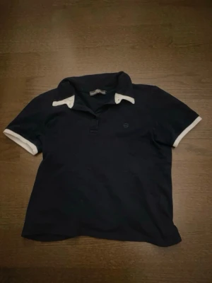 Marinblå pikétröja med vita detaljer - Klassisk marinblå pikétröja med vita detaljer på krage och ärmslut. Tröjan har korta ärmar och en liten diskret logga på bröstet. Perfekt till jeans eller shorts för en avslappnad look.