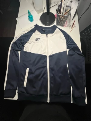 Blåvit träningsjacka från Umbro - Snygg blåvit träningsjacka från Umbro med dragkedja framtill och ribbade muddar. Jackan har Umbro-logga på bröstet och axlarna, samt vita detaljer längs ärmar och fickor. Perfekt för träning eller sportiga outfits.