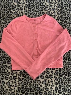 Rosa cropped kofta med knappar - Söt rosa cropped kofta med långa ärmar och knäppning framtill. Koftan har en mjuk, lite texturerad yta och rund halsringning. Perfekt att slänga över en topp för en chill look.