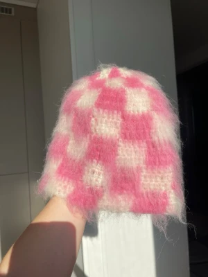 Rosa och vit fluffig rutig mössa - Supermjuk och fluffig mössa i rosa och vitt med rutigt mönster. Mössan har en avslappnad passform och är virkad i ett lurvigt material som ger en cozy vibe. Perfekt för dig som vill sticka ut med en färgglad accessoar i vår.