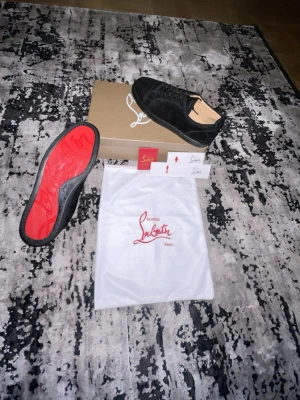Svarta Christian Louboutin sneakers - Snygga svarta sneakers från Christian Louboutin med klassisk röd sula och diskret logga. Skorna är låga och har en stilren design i mocka med svarta snören. Perfekta för dig som vill ha något exklusivt och trendigt.
