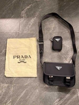 Svart axelväska - Snygg svart axelväska i nylon med justerbar axelrem och silverfärgade detaljer. Väskan har ett lock med spännen, ikonisk trekantslogga framtill och en liten avtagbar pouch med logga. Perfekt för dig som gillar stilrena och lyxiga accessoarer.