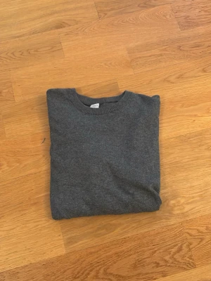 Mörkgrå stickad tröja  - En enkel och stilren mörkgrå stickad tröja med rund hals och långa ärmar. Tröjan har ribbade muddar vid ärmslut och nederkant. Perfekt basplagg för lager på lager och funkar till det mesta.