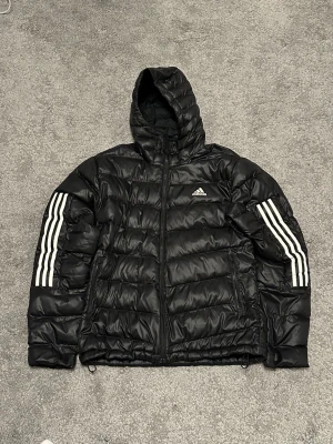 Svart pufferjacka från Adidas - Säljer en svart pufferjacka från Adidas med huva och dragkedja. Jackan har de klassiska vita Adidas-ränderna längs ärmarna och logga på bröstet. Tillverkad i glansigt syntetmaterial och har två sidofickor med dragkedja. Perfekt för kalla vinterdagar.
