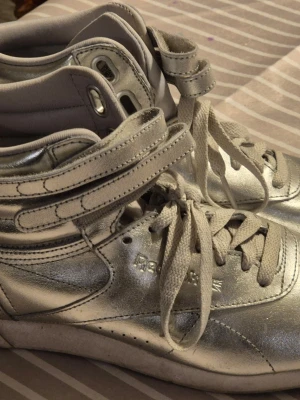 Silvriga Reebok high top sneakers - Säljer ett par riktigt snygga silvriga high top sneakers från Reebok. Skorna har metallic finish, dubbla kardborreband upptill och klassiska snörningar. Ovandelen är i skinn med vadderad insida för extra komfort. Perfekta för dig som vill sticka ut!