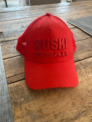 Röd truckerkeps från Huski Wear - Snygg röd truckerkeps från Huski Wear med broderad logga framtill och mesh på sidorna och bak för extra ventilation. Justerbar snapback baktill för perfekt passform. Perfekt accessoar för en sportig och avslappnad stil.