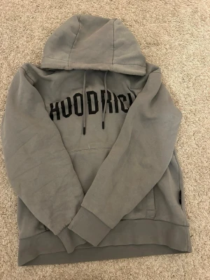 hoodrich hoodie - Säljer en grå hoodie från HOODRICH med stor svart logga över bröstet. Hoodien har en klassisk huva med dragsnören och en stor magficka framtill. Tillverkad i mjukt bomullsmaterial och har ribbade muddar vid ärmslut och nederkant.