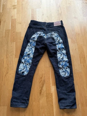 Japanska raw denim jeans med mönster - Unika mörkblå raw denim jeans från Evisu med ikoniskt handmålat blått och vitt mönster på bakstycket. Klassisk rak passform och detaljerad patch i läder med japanska tecken. Perfekta för dig som vill sticka ut med streetwear-vibe.