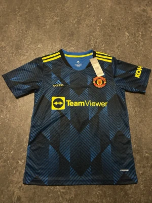 Manchester United bortatröja Adidas - Snygg Manchester United bortatröja från Adidas i mörkblått med svarta och blå mönster, gula detaljer och klubbmärke på bröstet. Tröjan har TeamViewer som sponsortryck och gula ränder på axlarna. Tillverkad i lätt och ventilerande material.