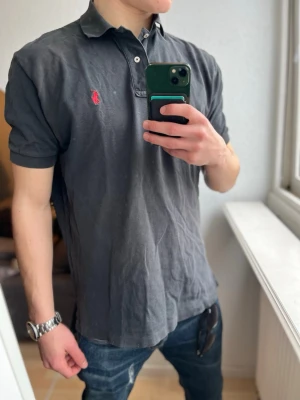 Svart piké från Polo Ralph Lauren - Klassisk svart pikétröja från Polo Ralph Lauren med korta ärmar och röd broderad logga på bröstet. Tröjan har krage, knappar vid halsen och är tillverkad i mjuk bomull. Perfekt till jeans eller shorts för en avslappnad look.