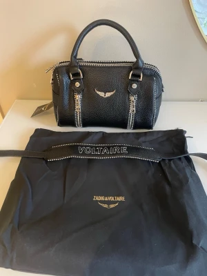 Svart handväska Zadig & Voltaire - Cool svart handväska från Zadig & Voltaire i strukturerat läder med silverfärgade detaljer och dragkedjor. Väskan har dubbla handtag, dekorativ logga med vingar framtill och medföljande dustbag. Perfekt för dig som gillar edgy och trendig stil.