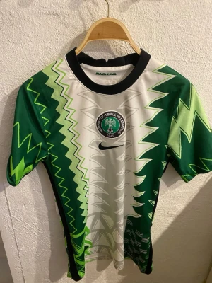 Nigeria fotbollströja Nike grön/vit - Säljer en Nigeria fotbollströja från Nike med ikoniskt grönt och vitt zigzag-mönster. Tröjan har korta ärmar, svart krage och Nigeria Football Federation-logga på bröstet. Tillverkad i lätt och ventilerande material, perfekt för träning eller match.