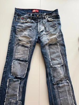 Blå jeans från Guess midjemått 37 - Säljer ett par blå jeans från Guess med kraftigt slitna detaljer och stora patchade hål framtill. Jeansen har raka ben, klassisk femficksmodell och tydliga fransiga kanter på lapparna. Perfekta för dig som gillar en edgy och trendig stil.