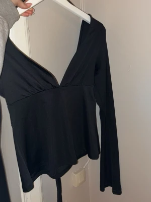 Svart omlottopp med v-ringning - Snygg svart omlottopp med djup v-ringning och långa ärmar. Toppen har en figurnära passform och knyts i midjan för en smickrande siluett. Perfekt att styla med jeans eller kjol för en trendig look.