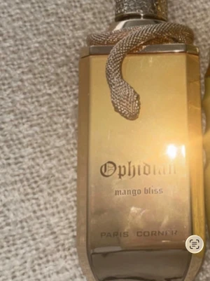 Ophidian Mango Bliss parfym - Lyxig parfymflaska i guld med en detaljerad orm runt halsen. Ophidian Mango Bliss från Paris Corner har en elegant, fyrkantig form och flaskan är tillverkad i glas med metalliska detaljer. Perfekt för dig som gillar unika och snygga parfymflaskor.