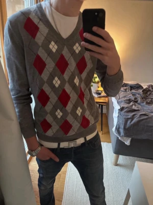 Grå argyle stickad tröja - Snygg grå stickad tröja med klassiskt argyle-mönster i rött, vitt och mörkgrått. Tröjan har V-ringning och långa ärmar, perfekt för lager på lager. Passar till både jeans och chinos för en stilren look.