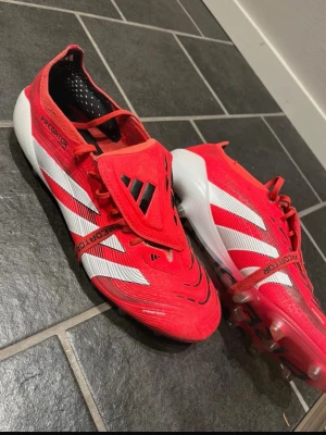 Adidas Predator röda fotbollsskor - Säljer ett par Adidas Predator fotbollsskor i rött med vita och svarta detaljer. Skorna har klassisk snörning, täckt plös och mönstrad ovandel för extra grepp. Yttersulan har fasta dobbar för spel på gräs. Perfekta för dig som vill dominera på planen.