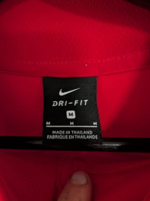 Röd Nike Dri-Fit träningströja - Snygg röd träningströja från Nike med Dri-Fit material som andas. Tröjan har halv dragkedja framtill, vit Nike-logga på bröstet och vita detaljer över bröstet. Perfekt för träning eller uppvärmning.