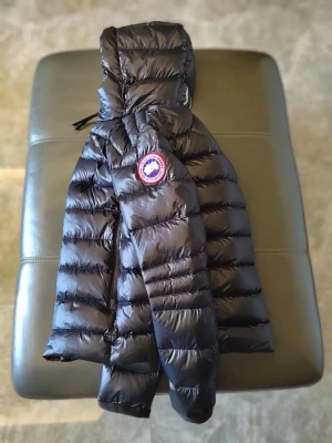 Svart dunjacka från Canada Goose - Snygg svart dunjacka från Canada Goose med glansig finish och klassisk logotyp på ärmen. Jackan har huva, dragkedja framtill och quiltad design som håller värmen under kalla vinterdagar. Perfekt för dig som vill ha både stil och funktion.