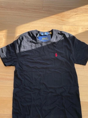 Svart Ralph Lauren T-shirt  - 👕 Märke: Polo Ralph Lauren 📏 Storlek: S 🎨 Färg: Svart med röd logga ✨ ☀️ Stilren och snygg t-shirt från Polo Ralph Lauren – perfekt för sommaren och enkel att matcha med både shorts och jeans. Tillverkad i mjuk och bekväm bomull med klassisk passform och ikonisk röd logga på bröstet.  ✨ Skick: Helt ny med tags kvar 🧼 Aldrig använd 👌 Tidlös modell som passar till allt