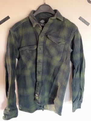 Grönrutig flanellskjorta från Dickies - Snygg grön och mörkblå rutig skjorta från Dickies med klassisk krage och två bröstfickor med knapp. Skjortan har långa ärmar och knäppning framtill. Perfekt för en avslappnad och cool stil.