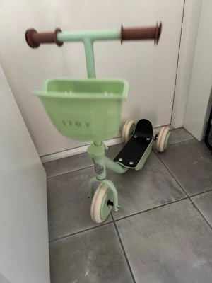 Mintgrön sparkcykel med korg - Säljer en ljusgrön sparkcykel för barn från Stoy. Den har fyra hjul, brunt styre med greppvänliga handtag och en praktisk korg framtill. Cykeln är tillverkad i metall och plast, och har en svart fotplatta. Perfekt för små äventyr inomhus eller utomhus. Detta är en använd för att vi köpte en men råkade beställa tre till så de andra tre är inpackade i original paketen helt orörda.