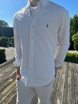 Vit skjorta från Ralph Lauren - (MODDELEN ÄR 185 OCH VÄGER 75)Klassisk vit skjorta från Ralph Lauren med broderad logga på bröstet. Skjortan har button-down krage, långa ärmar och stängs med vita knappar. Perfekt för en clean och stilren look.