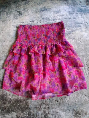 Blommig volangkjol från BikBok - Söt kortkjol från BikBok i blommigt mönster med rosa, lila och röda toner. Kjolen har smock i midjan och flera lager volanger för en flowy känsla. Perfekt för dig som gillar färg och vill sticka ut lite extra.