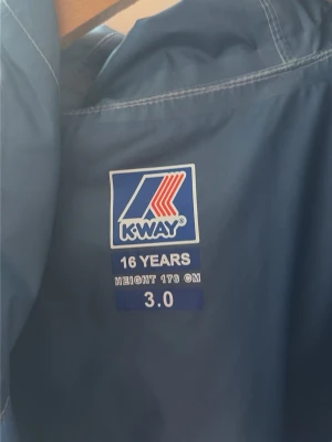 Blå vindjacka från K-Way - Snygg blå vindjacka från K-Way med huva och dragkedja med färgglada detaljer. Jackan är lätt och perfekt för blåsiga dagar. Klassisk logga på bröstet och smidig passform. Materialet är tunt och vattenavvisande.
