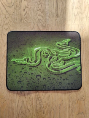 Razer musmatta 270x215 - Säljer en rektangulär musmatta från Razer med svart kant och en stor grön Razer-logga på ovansidan. Undersidan är helt grön och har en texturerad yta för bra grepp. Perfekt för gaming eller datorarbete.