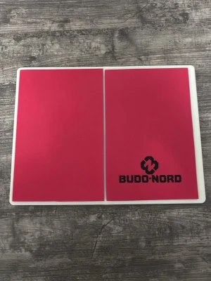 Rosa plastbrädor från Budo-Nord - Två rektangulära rosa plastbrädor från Budo-Nord med vit kant. Ena brädan har Budo-Nord-loggan tryckt i svart. Perfekta för träning eller kampsport, tillverkade i tålig plast. Denna är den näst hårdaste brädan man kan köpa och jag betala 400 kr på detta