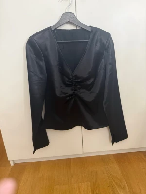 Svart satinblus med v-ringning - Stilren svart blus i glansigt satinmaterial med djup v-ringning framtill. Långa ärmar och figurnära passform ger en elegant look. Perfekt för dig som gillar trendiga och klassiska plagg.