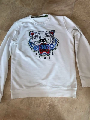 Vit sweatshirt från Kenzo med tigertryck - Säljer en vit sweatshirt från Kenzo med det ikoniska broderade tigerhuvudet och färgglad text på bröstet. Tröjan har rund halsringning, långa ärmar och är gjord i mjukt bomullsmaterial. Perfekt för dig som gillar streetwear och vill sticka ut.