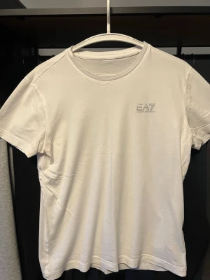 Vit EA7 t-shirt - Snygg vit t-shirt från EA7 Emporio Armani med diskret logga på bröstet. Klassisk rund halsringning och korta ärmar. Tillverkad i mjuk bomull som känns skön mot huden. Perfekt för en clean och stilren look.