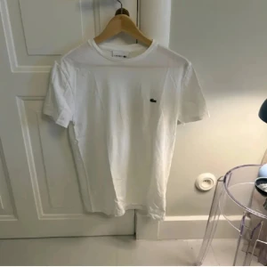 Vit t-shirt från Lacoste - Säljer en klassisk vit t-shirt från Lacoste med den ikoniska krokodilloggan på bröstet. T-shirten har en normal passform och är tillverkad i mjuk bomull. Perfekt för en stilren och avslappnad look.