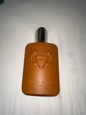 Parfums de Marly Althair - Säljer en exklusiv parfym från Parfums de Marly i en matt orange flaska med silverfärgat lock. Flaskan har ett elegant reliefmönster med två hästar och texten '1743' samt 'PARFUMS de MARLY PARIS' ingraverat framtill. Perfekt för dig som gillar lyxiga dofter. Runt 80% kvar i flaskan