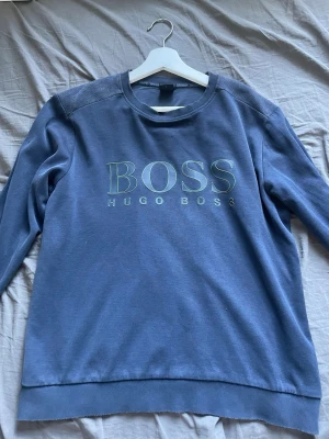 Blå sweatshirt från Hugo Boss - Blå sweatshirt från Hugo Boss med stort BOSS-tryck på bröstet. Storleken är slim fit L men den sitter mer som en M. Perfekt för en avslappnad och stilren look.