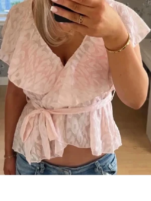 Ljusrosa omlottblus med knytband - EJ MIN BILD PÅ FÖRSTA! Söt ljusrosa omlottblus med diskret mönster och volangig v-ringning. Blusen har korta ärmar och knyts i midjan med ett matchande band. Tunt, lätt material som ger en luftig känsla. Perfekt för en trendig och feminin look.