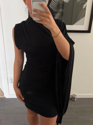 Svart oneshoulder kortklänning - Zara strl S - Snygg svart kortklänning med unik oneshoulder-design och draperad detalj som hänger ner från ena axeln. Klänningen har en tight passform och är perfekt för dig som vill sticka ut. Materialet känns mjukt och följsamt mot kroppen. Snygg till finare tillfälle - den perfekta lilla svarta klänningen!