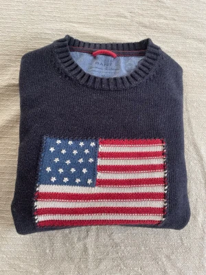 Mörkblå stickad tröja från Gant - Snygg mörkblå stickad tröja från Gant med amerikansk flagga framtill. Tröjan har rund halsringning och är tillverkad i 100% bomull. Perfekt för dig som gillar klassisk preppy stil och vill ha något som sticker ut.